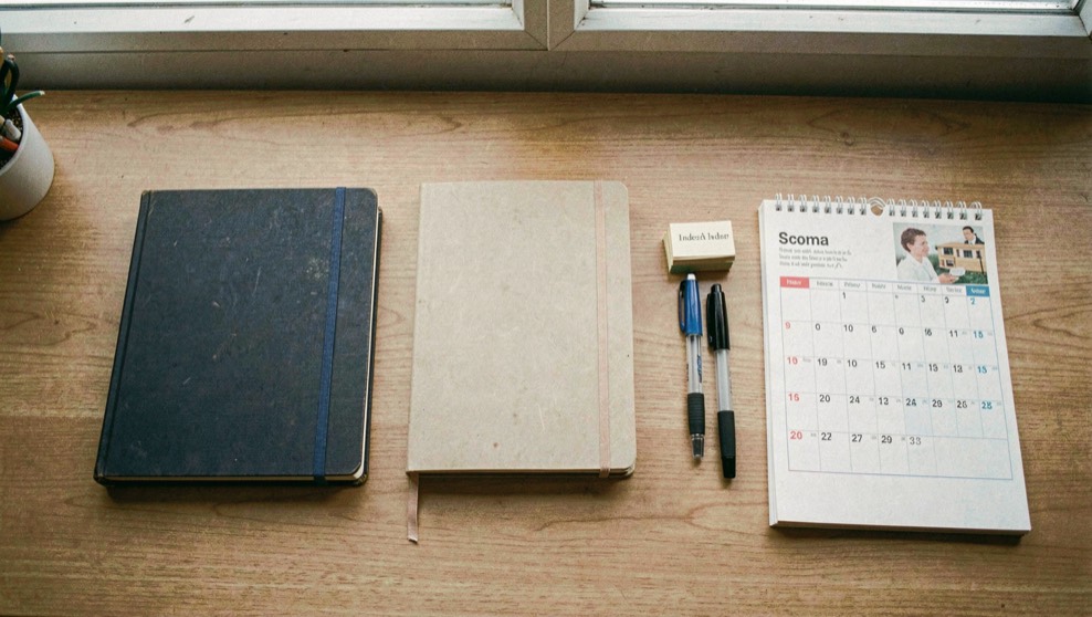 An overhead view of a plain wooden desk with three notebooks of different sizes, a stack of index cards, two pens and a paper calendar arranged in a row, even daylight from a window.