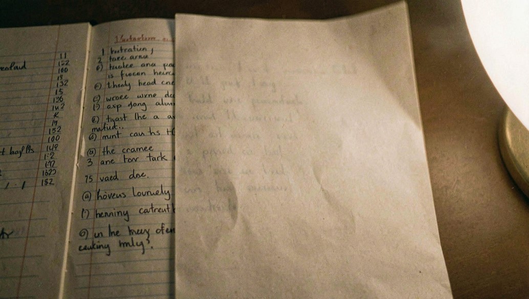 A hand writing short questions down the narrow left margin of a filled notebook page, the wide column already covered by a folded sheet of paper, evening lamp light from the right.